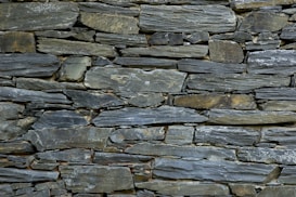 A tightly fitted stone wall made of irregularly shaped, flat pieces of slate arranged horizontally. The color palette includes various shades of gray, with subtle hints of green and brown, and the surface texture is rugged and natural.