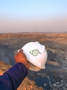 A hand holding a white hard hat with a green symbol, overlooking a large, open quarry or mining site. The landscape consists of rocky terrain with an expansive view into the horizon, where the sky meets the earth, filled with a light blue and orange gradient from the setting or rising sun.