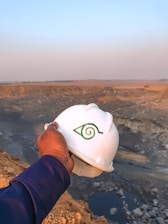 A hand holding a white hard hat with a green symbol, overlooking a large, open quarry or mining site. The landscape consists of rocky terrain with an expansive view into the horizon, where the sky meets the earth, filled with a light blue and orange gradient from the setting or rising sun.