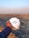 A hand holding a white hard hat with a green symbol, overlooking a large, open quarry or mining site. The landscape consists of rocky terrain with an expansive view into the horizon, where the sky meets the earth, filled with a light blue and orange gradient from the setting or rising sun.