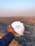 A hand holding a white hard hat with a green symbol, overlooking a large, open quarry or mining site. The landscape consists of rocky terrain with an expansive view into the horizon, where the sky meets the earth, filled with a light blue and orange gradient from the setting or rising sun.