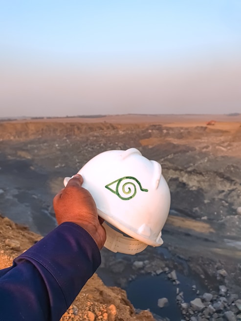 A hand holding a white hard hat with a green symbol, overlooking a large, open quarry or mining site. The landscape consists of rocky terrain with an expansive view into the horizon, where the sky meets the earth, filled with a light blue and orange gradient from the setting or rising sun.