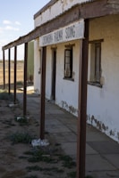 An old, weathered building with a sign that reads CORNUCURRA RAILWAY SIDING. The structure has peeling paint on its walls, rusted metal support beams, and broken windows. The ground is a mix of dirt and patches of grass, and the scene appears to be located in a desolate, arid area under a clear sky.
