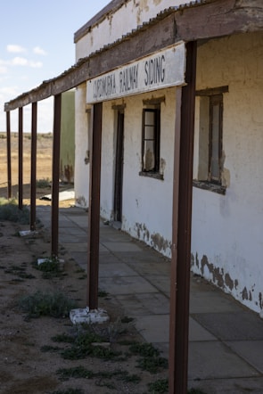 An old, weathered building with a sign that reads CORNUCURRA RAILWAY SIDING. The structure has peeling paint on its walls, rusted metal support beams, and broken windows. The ground is a mix of dirt and patches of grass, and the scene appears to be located in a desolate, arid area under a clear sky.
