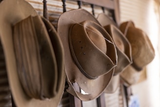 Several brown, worn cowboy hats are hung in a row on a wooden rack. The hats appear weathered, with noticeable creases and signs of age. The background includes a corrugated metal surface, adding to a rustic and vintage atmosphere.