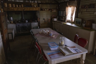A rustic, dimly lit kitchen features a large wooden table with several chairs, some with red seats. Various jars and containers sit on the table, alongside a book and pens. A vintage stove is situated along the back wall, surrounded by aged metal and wooden surfaces. The walls and ceiling are made from corrugated metal, giving the space a quaint, old-fashioned charm. A small window with floral curtains allows some light to enter, casting soft shadows across the room. Kitchen implements and pictures adorn the walls.