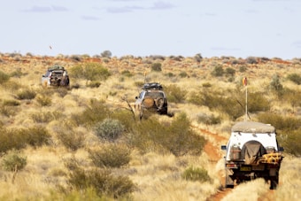 A rugged landscape featuring a dirt trail leading through a vast desert area, with three off-road vehicles traveling in a convoy. Sparse vegetation covers the arid terrain, and the sky is clear with a few clouds. Each vehicle carries camping gear, highlighting the adventurous nature of the journey.