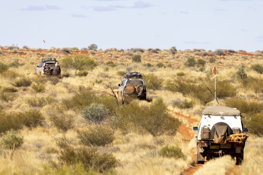 A rugged landscape featuring a dirt trail leading through a vast desert area, with three off-road vehicles traveling in a convoy. Sparse vegetation covers the arid terrain, and the sky is clear with a few clouds. Each vehicle carries camping gear, highlighting the adventurous nature of the journey.
