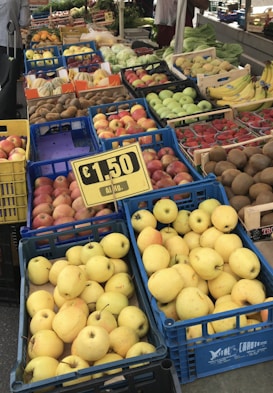 Various fruits such as apples, kiwis, bananas, and strawberries are displayed in blue crates at a market stall. A sign indicates a price of €1.50 per kilogram. The background shows additional produce and green leafy vegetables.