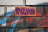 A brick wall features graffiti-style text that reads 'FUTURE IS CREATIVE' in bold purple letters against an orange background. Surrounding the text are vibrant spray-painted patterns in shades of blue, red, and white, providing a colorful and artistic backdrop.