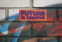 A brick wall features graffiti-style text that reads 'FUTURE IS CREATIVE' in bold purple letters against an orange background. Surrounding the text are vibrant spray-painted patterns in shades of blue, red, and white, providing a colorful and artistic backdrop.