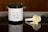 A container labeled 'protein' with a design of a muscular arm is placed on a reflective surface. Next to it, a white scoop with protein powder is partially filled and some powder is spilled on the surface.