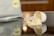 A white measuring scoop filled with a light beige powder rests on a reflective surface, with some powder spilled in front of it. In the background, a package labeled 'Vanilla' is partially visible.