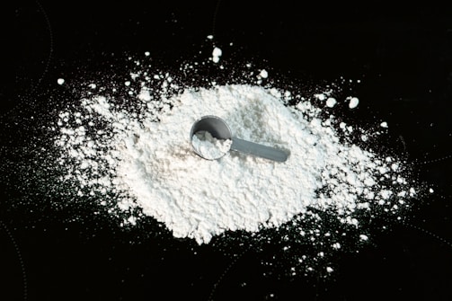 A pile of white powder is spread across a dark surface with a small metal scoop resting on top of the powder. The powder appears finely textured and is scattered around the scoop.