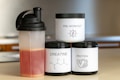There are three supplement containers and a shaker bottle. One container is labeled 'PRE-WORKOUT' with a lightning bolt symbol, another is labeled 'CREATINE' with a chemical structure diagram, and the third is labeled 'PROTEIN' with an icon of a flexing arm. The shaker bottle contains a red liquid.