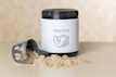 A container of protein powder with a white label displaying the word 'PROTEIN' and an icon of a flexed arm. The container has a black lid and is placed on a flat surface. In front of it, there is a scoop partially filled with the powder, some of which is spilled on the surface.