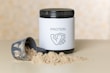 A container of protein powder with a white label displaying the word 'PROTEIN' and an icon of a flexed arm. The container has a black lid and is placed on a flat surface. In front of it, there is a scoop partially filled with the powder, some of which is spilled on the surface.