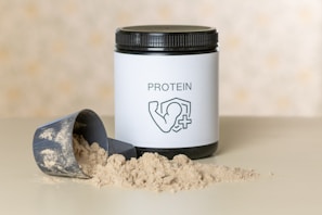 A container of protein powder with a white label displaying the word 'PROTEIN' and an icon of a flexed arm. The container has a black lid and is placed on a flat surface. In front of it, there is a scoop partially filled with the powder, some of which is spilled on the surface.