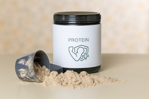A container of protein powder with a white label displaying the word 'PROTEIN' and an icon of a flexed arm. The container has a black lid and is placed on a flat surface. In front of it, there is a scoop partially filled with the powder, some of which is spilled on the surface.