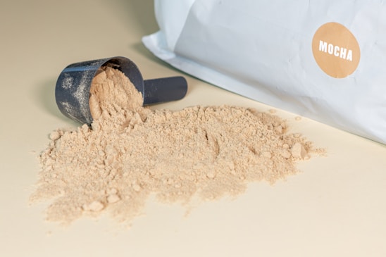 A black scoop filled with beige powder spills onto a surface. A large white bag with a circular mocha-colored label rests beside it.