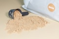 A black scoop filled with beige powder spills onto a surface. A large white bag with a circular mocha-colored label rests beside it.