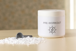 A container labeled 'PRE-WORKOUT' with a lightning bolt symbol sits on a surface. Beside it, there is a small pile of white powder and a grey scoop, suggesting a supplement or powder for fitness purposes.