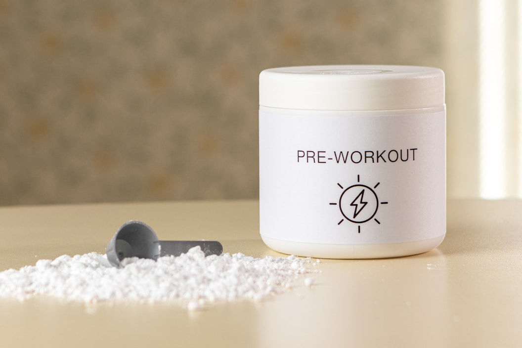 A container labeled 'PRE-WORKOUT' with a lightning bolt symbol sits on a surface. Beside it, there is a small pile of white powder and a grey scoop, suggesting a supplement or powder for fitness purposes.