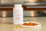 A white bottle labeled Omega-3 with a fish icon is placed on a countertop. Beside it, there are several translucent yellow capsules and a small white plate containing slices of raw salmon. The background features a blurred kitchen setting with warm lighting.