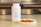 A white bottle labeled Omega-3 with a fish icon is placed on a countertop. Beside it, there are several translucent yellow capsules and a small white plate containing slices of raw salmon. The background features a blurred kitchen setting with warm lighting.