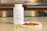 A white bottle labeled Omega-3 with a fish icon is placed on a countertop. Beside it, there are several translucent yellow capsules and a small white plate containing slices of raw salmon. The background features a blurred kitchen setting with warm lighting.