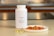 A white bottle labeled Omega-3 with a fish icon is placed on a countertop. Beside it, there are several translucent yellow capsules and a small white plate containing slices of raw salmon. The background features a blurred kitchen setting with warm lighting.