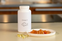 A white bottle labeled Omega-3 with a fish icon is placed on a countertop. Beside it, there are several translucent yellow capsules and a small white plate containing slices of raw salmon. The background features a blurred kitchen setting with warm lighting.