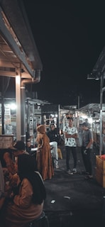 A bustling night market scene with several people standing and engaging in conversation. The surroundings include stalls with various signs and advertisements. The lighting creates a warm, inviting atmosphere, with a contrast between the light and dark areas.