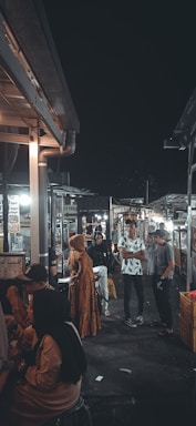 A bustling night market scene with several people standing and engaging in conversation. The surroundings include stalls with various signs and advertisements. The lighting creates a warm, inviting atmosphere, with a contrast between the light and dark areas.