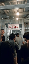 A group of people are standing in line inside a metal-roofed structure. There is a sign hanging above that says 'KASIR' in red, which suggests a cashier or payment area. The atmosphere is busy, with individuals appearing to be waiting or queuing.