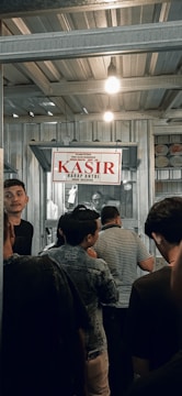 A group of people are standing in line inside a metal-roofed structure. There is a sign hanging above that says 'KASIR' in red, which suggests a cashier or payment area. The atmosphere is busy, with individuals appearing to be waiting or queuing.