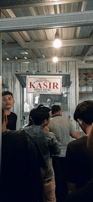 A group of people are standing in line inside a metal-roofed structure. There is a sign hanging above that says 'KASIR' in red, which suggests a cashier or payment area. The atmosphere is busy, with individuals appearing to be waiting or queuing.