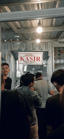 A group of people are standing in line inside a metal-roofed structure. There is a sign hanging above that says 'KASIR' in red, which suggests a cashier or payment area. The atmosphere is busy, with individuals appearing to be waiting or queuing.