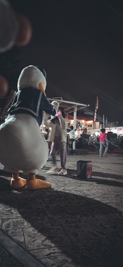 A large costumed character resembling a duck interacts with a person in an outdoor setting at night. The scene appears to be festive, with other people in the background and decorative lights. The atmosphere is lively, with a few individuals walking around.