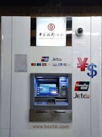 An ATM machine associated with the Bank of China (Hong Kong) featuring logos of various payment networks such as UnionPay, Jetco, Visa, and Plus. Above the machine, a surveillance camera is visible. The design is predominantly white with blue and metallic elements.