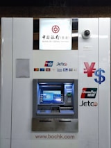 An ATM machine associated with the Bank of China (Hong Kong) featuring logos of various payment networks such as UnionPay, Jetco, Visa, and Plus. Above the machine, a surveillance camera is visible. The design is predominantly white with blue and metallic elements.