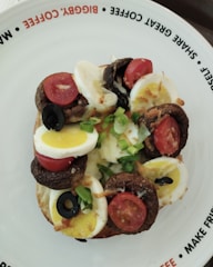 A toasted bagel topped with slices of hard-boiled egg, cherry tomatoes, black olives, sautéed mushrooms, and chopped green onions. The plate features playful text on the rim promoting sharing and enjoying coffee together.