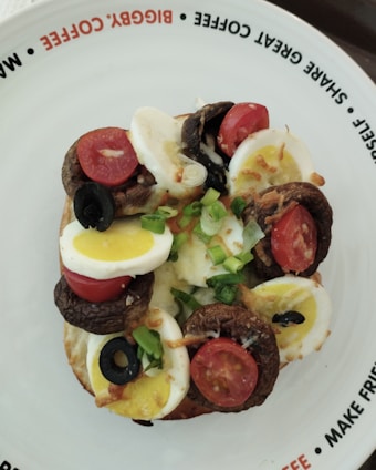 A toasted bagel topped with slices of hard-boiled egg, cherry tomatoes, black olives, sautéed mushrooms, and chopped green onions. The plate features playful text on the rim promoting sharing and enjoying coffee together.