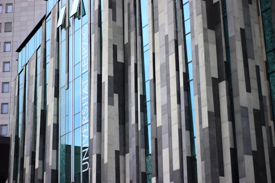 A modern building facade with a series of vertical panels and large glass windows. The panels display a pattern of alternating dark and light grey tones, interspersed with narrow blue-tinted window panes. The reflection of the sky enhances the glass surfaces. There is text visible on one of the panels.