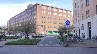A city street scene featuring a designated bicycle lane surrounded by green spaces and several trees. A multi-story brick building is in the background with many windows, along with another building on the right. Numerous bicycles are parked around the area. There are road signs indicating a bicycle path.