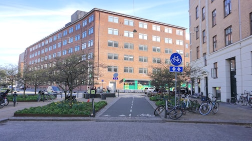 A city street scene featuring a designated bicycle lane surrounded by green spaces and several trees. A multi-story brick building is in the background with many windows, along with another building on the right. Numerous bicycles are parked around the area. There are road signs indicating a bicycle path.