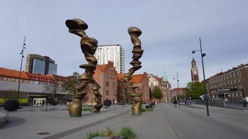 A modern urban area features a large, abstract bronze sculpture with twisting forms in the foreground. Surrounding buildings vary in architectural style, including red-roofed and brick structures, with a tall clock tower and a high-rise in the distance. The street is lined with streetlights, a few trees, and sparse pedestrian and vehicular activity.