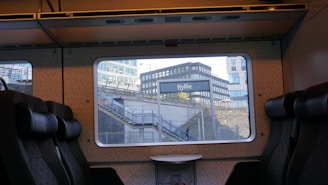 View from the interior of a train, showing empty seats and a table. Outside the window is a train station with the sign 'Hyllie'. Modern buildings and a staircase are visible in the background. A single person is walking under the staircase.