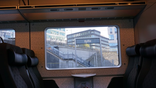 View from the interior of a train, showing empty seats and a table. Outside the window is a train station with the sign 'Hyllie'. Modern buildings and a staircase are visible in the background. A single person is walking under the staircase.
