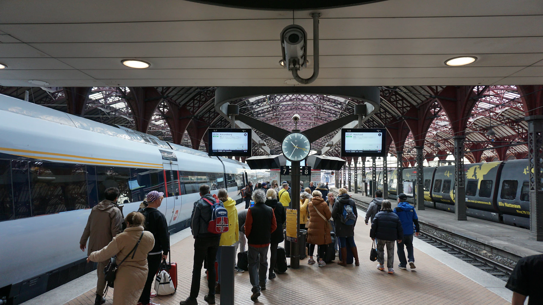 Passengers with bags wait beside trains at a Copenhagen station platform.